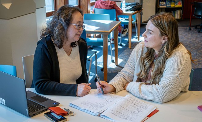 A professor and a student meet in a lounge area with a laptop and a notebook full of notes open on the table.