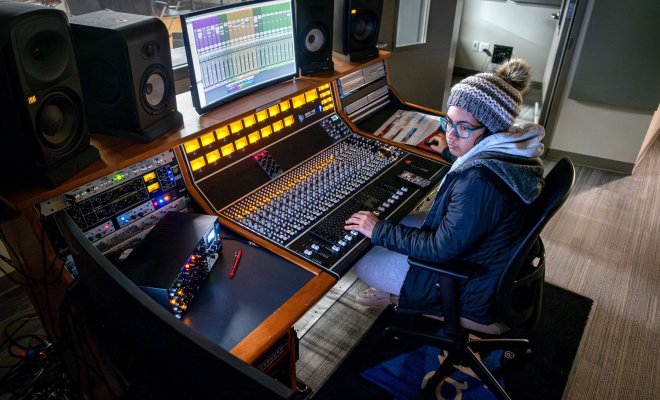 A student sits in front of a large mixing board in a professional-grade recording studio, the performance space visible through a window in front of her.