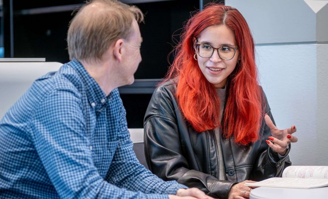 A red-haired student in a leather jacket converses with a faculty member in a blue and white checkered shirt.