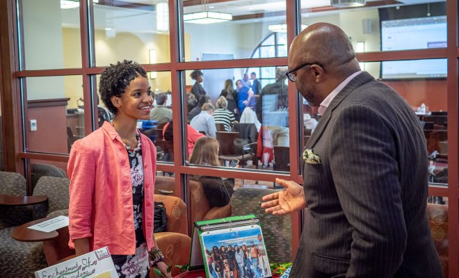 Student in business attire stands behind a table at a pitch competition, speaking to a business professional