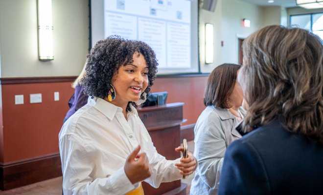 A student has a dialogue with a professor in a large meeting area with her sales pitch slides displayed on the large screen behind her.
