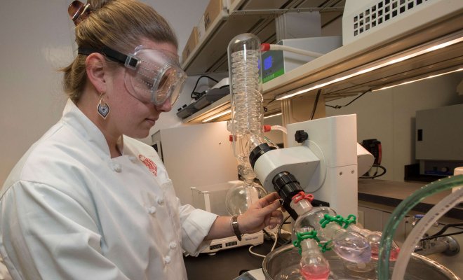 Student in chef's jacket wearing safety goggles working in the food lab.
