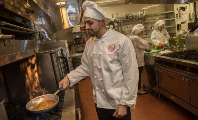 A student in a white chef's jacket and hat cooks food in a frying pan in a combination laboratory and kitchen.