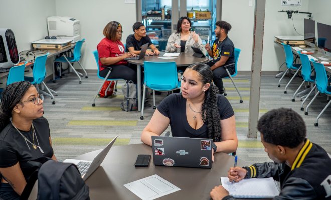 Three students gather around a small table to brainstorm, while a group of four students at another table in the background do the same.
