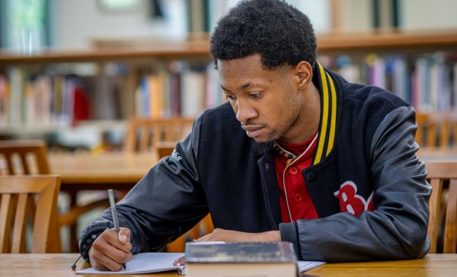 A student in a Bloomfield varsity jacket sits at a table in the library and writes notes in a notebook.