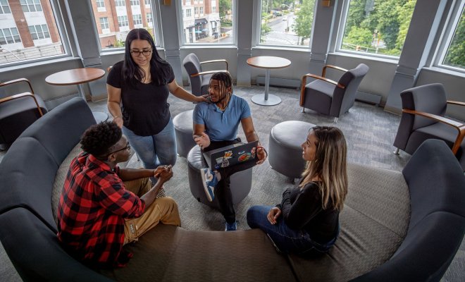 Four students gathered in a lounge talking with one another. The curved wall lined with windows behind them shows a view of downtown Bloomfield.