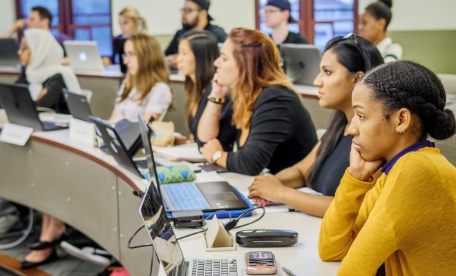 Two rows of students in a classroom with the desks arranged in tiers to make an amphitheater-style presentation space.