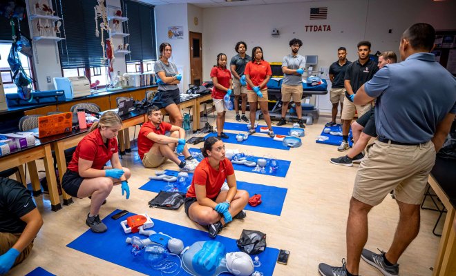 Students in a classroom with mats laid out on the floor and an assortment of training equipment arrayed on each mat.