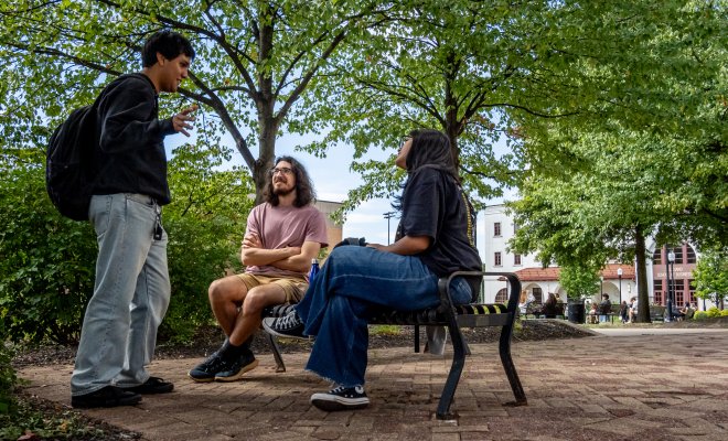 Three students are on campus, two are sitting on a park bench while one is standing as they are conversating with trees behind them. 