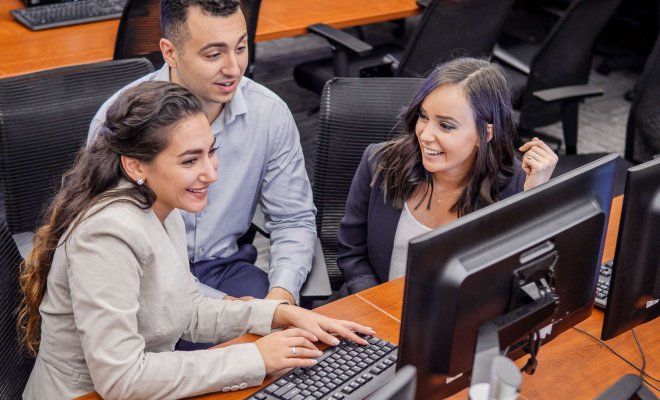 Three students in businesswear collaborate on a project in front of a multi-monitor setup.