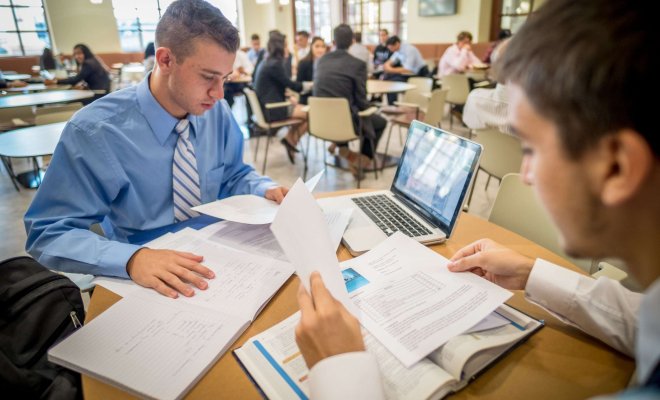 Two students in button-down shirts and ties at a cafe table with books and notes spread out and a laptop.