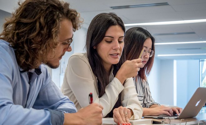 Two students in conversation at a table while a third student works on a laptop.