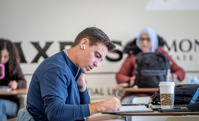Student wearing earbuds, siting at a table and writing in notebook.