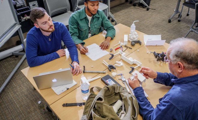 Two students and a professor sit at a work bench with a laptop and an assortment of 3D printed parts.