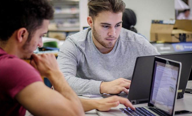 Two students sit at a table in the cafeteria intently working on their laptops.