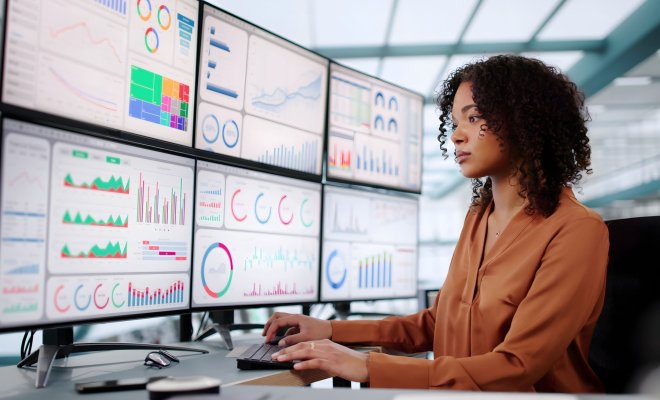 Woman in office sits at desk with six monitors displaying assorted charts and graphs