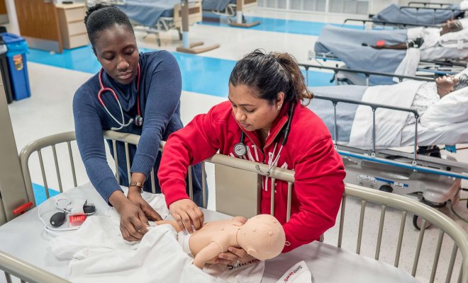 Two nursing students in a hospital-like classroom reach into a crib to practice procedures on a baby test dummy