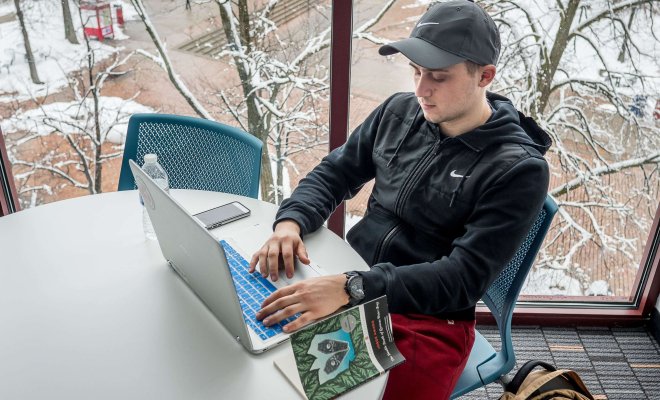 A student uses a laptop in a lounge in front of a large window overlooking the main quad in winter