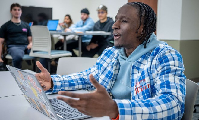 In a classroom, a student with an open laptop gestures while speaking.