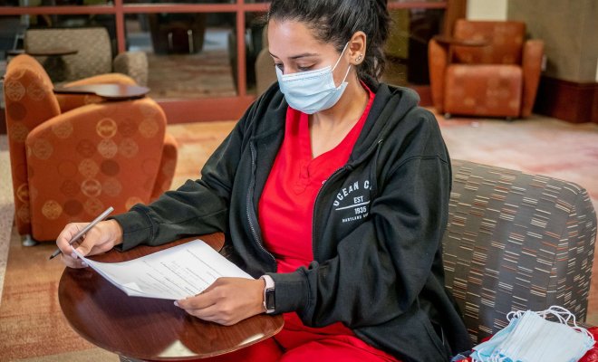 A student in red scrubs and a surgical mask completes her homework in a waiting room