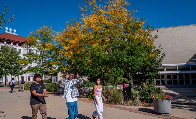 Three students are walking across campus, one student is giving a thumbs up to the camera.