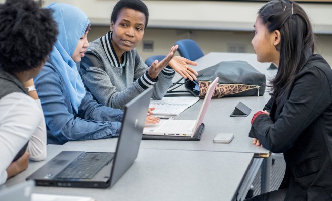 Graduate students at a table discussing over laptops and hand gestures.