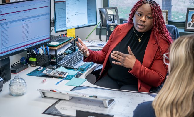 A professional staff member in a red blazer speaks with a student across a desk in an office.