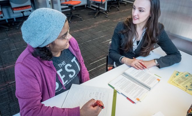 Student and professor sat down at a table discussing with pens and papers in front of them.