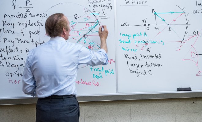 Professor in front of a whiteboard using a marker to write on board.