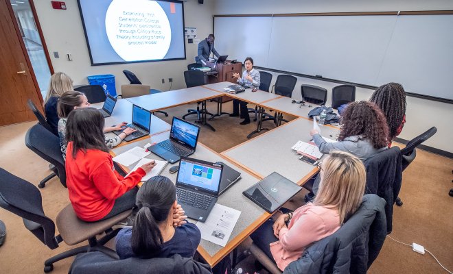 Student in a square shape desk wise at their laptops listening to their professor give a lecture.