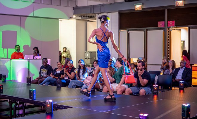 Student is walking a stage wearing a blue dress with students in the crowd looking.