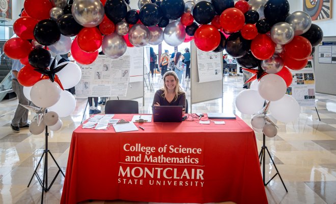 Table with the words College of Science and Mathematics on the red tablecloth, balloons wrapped around a woman smiling with a laptop in front of her.