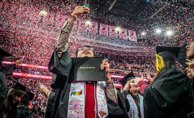 Student at commencement holding diploma holder and raising phone in the air as confetti falls