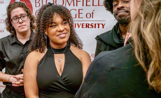 Ciara Donaldson, a 2024 Bloomfield College of Montclair State University graduate, stands at the center of the photo wearing a sleeveless black dress and a heart-shaped necklace, engaged with people gathered around her. Other individuals stand nearby, and a Bloomfield College of Montclair State University backdrop fills the background.