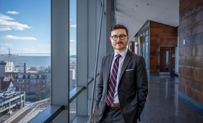 Student wearing suit and glasses faces the camera in long hallway with floor-to-ceiling windows