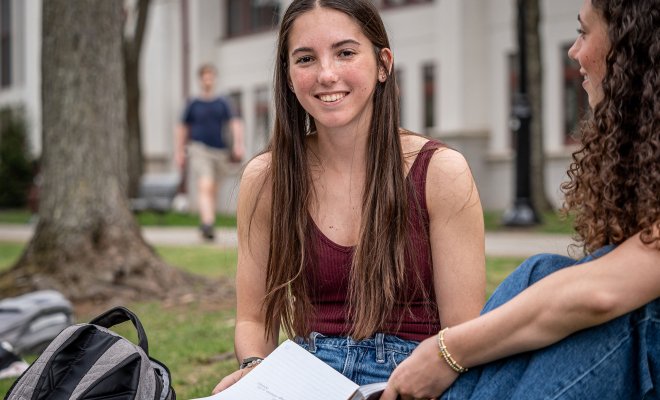 Dana Brzezinski sits on the grass at Montclair State University, holding an open notebook and talking with a friend, with campus buildings and a tree visible in the background.