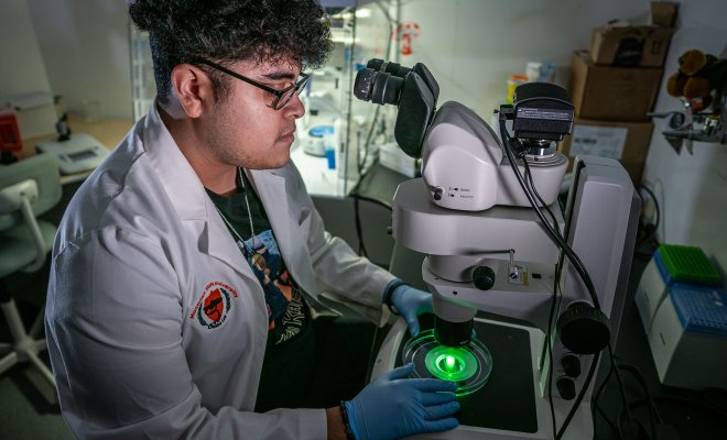 Melvin Rodriguez, wearing a white lab coat with a Montclair State University patch and blue gloves, sits at a fluorescent microscope in a dim lab. He looks intently through the eyepiece while holding a petri dish glowing bright green under the microscope lens, surrounded by scientific equipment and incubators in the background.