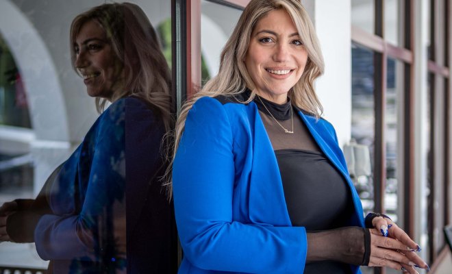 Ashley Crawford, in a black dress with bright blue blazer, stands in front of a building’s windows.
