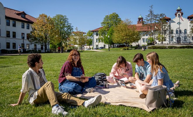 Students sit together on a blanket on a large grassy lawn at Montclair State University, talking and studying with notebooks and a laptop open, while academic buildings with red-tiled roofs and a clear blue sky frame the background.