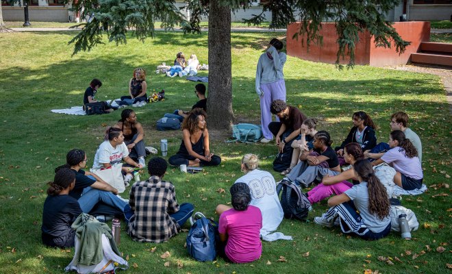 Students sitting outside on the grass and under a tree. They are sitting in a circle