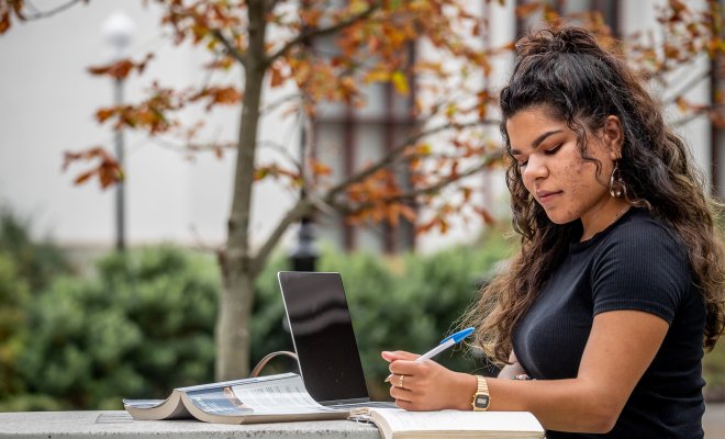 Female student at a table writing down notes in front of a laptop.