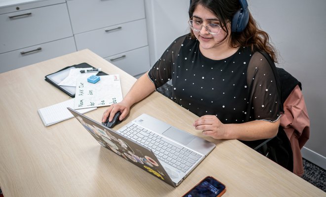 Student working on their computer with headphones on. They have written notes one their rightside.