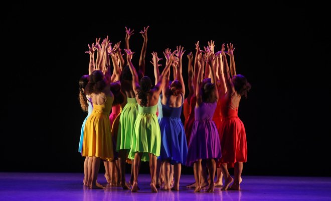 Performers on stage with their hands raised wearing different colored dresses.