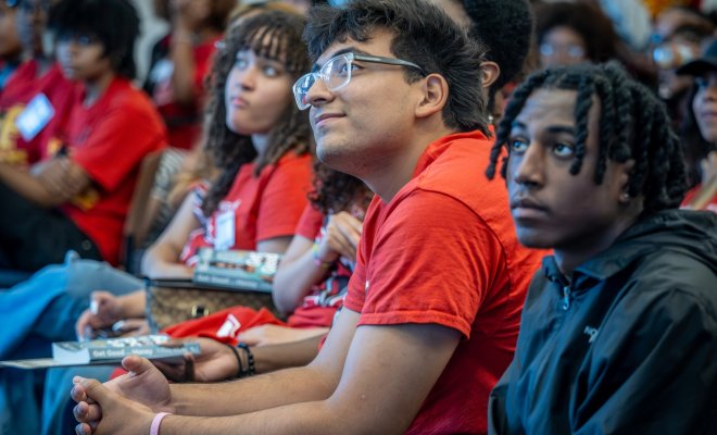 Franco Leon, wearing a red EOF T-shirt sits with hands clasped, smiling and looking up attentively during a conference session.