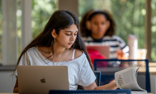 A student turns the pages to a book while working on her laptop in the library. A student wearing headphones sits behind her.
