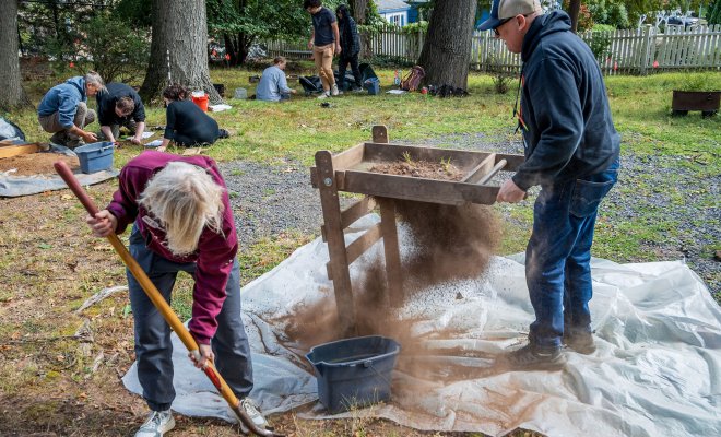 At a backyard archaeological dig, a woman digs a hole in the ground while a man uses a soil screener and others work in the background.