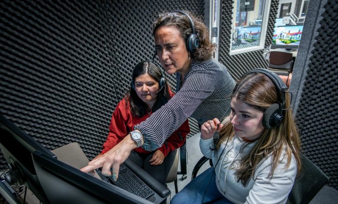 A professor with two students wearing headphones conversing while pointing to a computer screen.