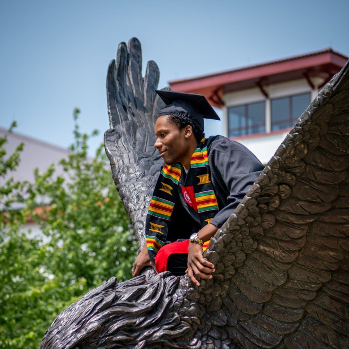 Male student in cap and gown with EOF stole poses for a photo on top of the hawk statue on campus