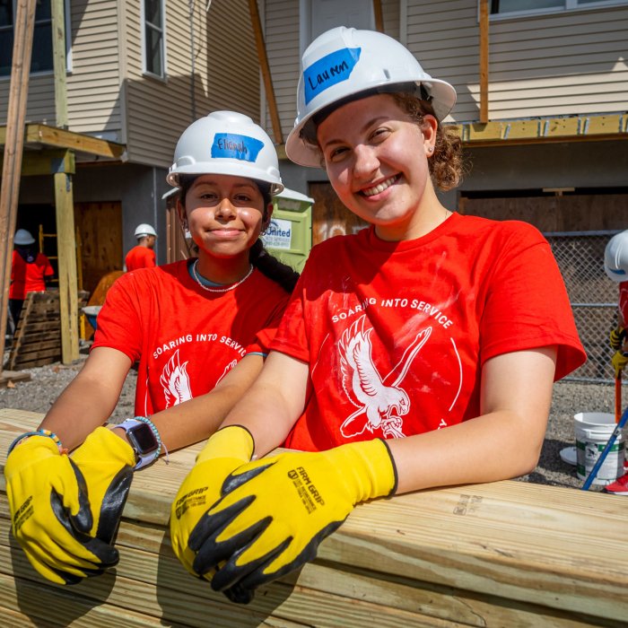 Two women students in red volunteer shirts take a break to pose for a photo while working to build houses with Habitat for Humanity