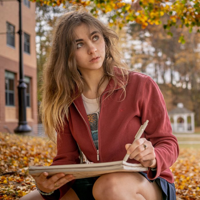 A student sits on campus in the fall sketching trees